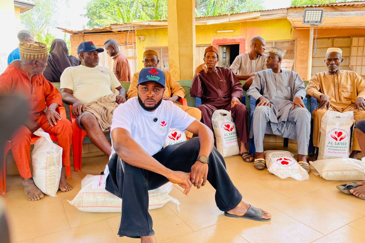 Older men seated with Al Sa'eed Humanitarian Foundation relief bags during a community distribution, with a Foundation volunteer.