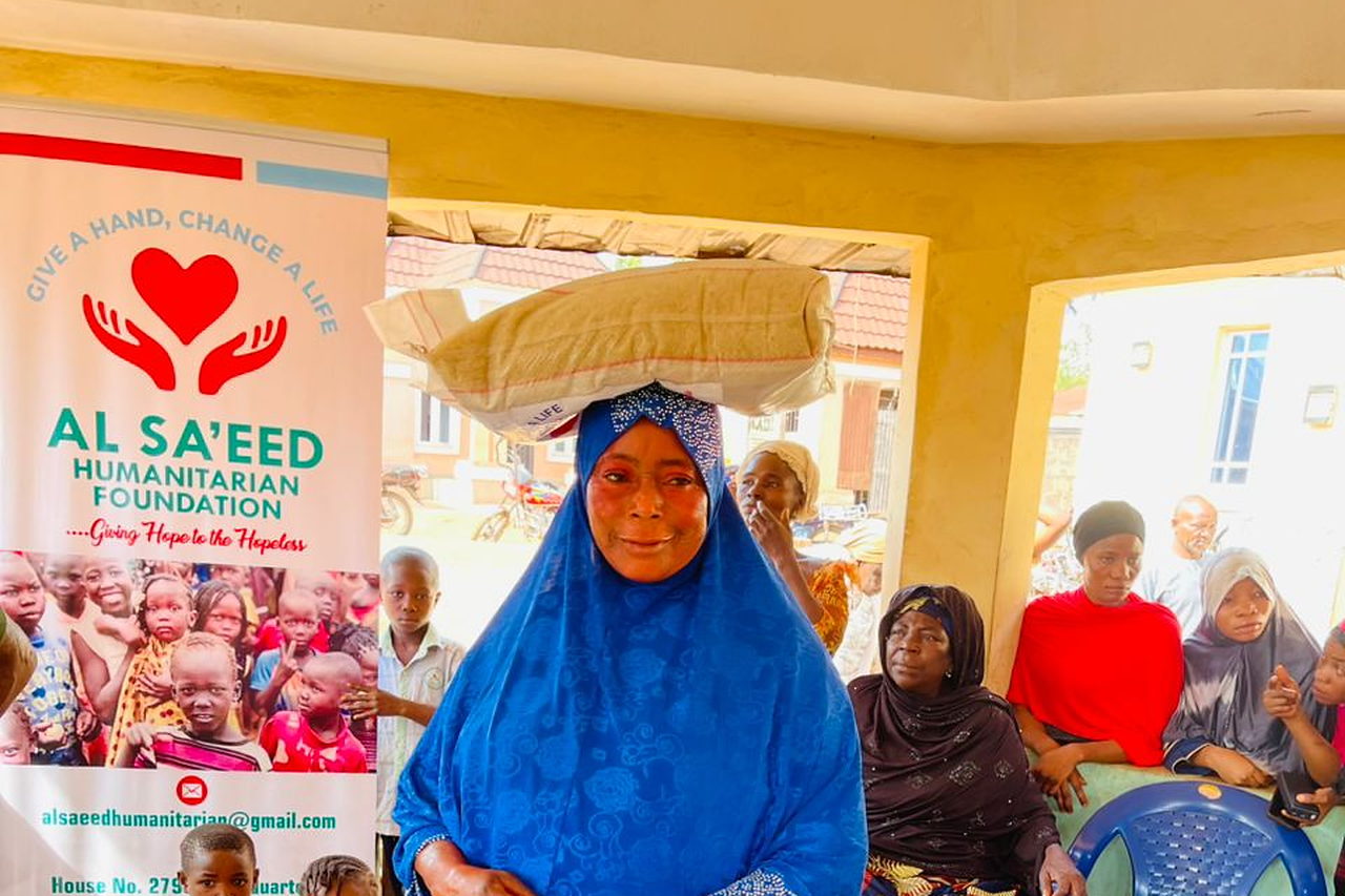 A woman carrying a bag of relief supplies on her head beside an Al Sa'eed Humanitarian Foundation banner.